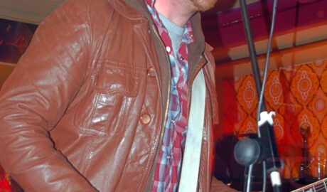 Low view of a man with short dark hair and beard, wearing a brown leather jacket over a brown hoodie, over a red and blue plaid shirt, over a grey t-shirt, playing a red electric guitar, while singing into a microphone on a stand. Orange paisley wallpaper covers the wall in the background, with orange and purple coping at the top, and pale yellow ceiling.