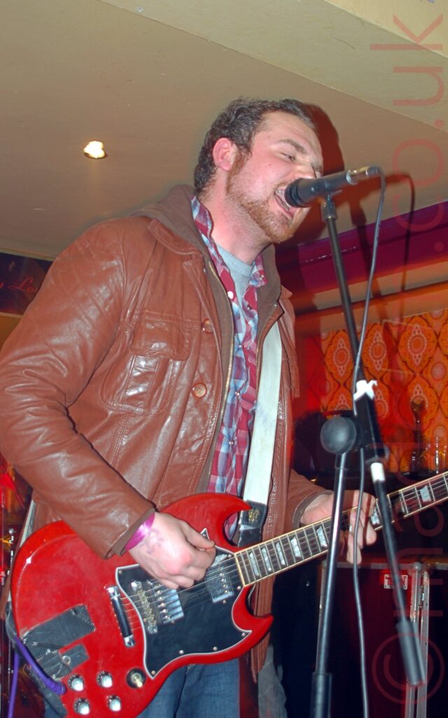 Low view of a man with short dark hair and beard, wearing a brown leather jacket over a brown hoodie, over a red and blue plaid shirt, over a grey t-shirt, playing a red electric guitar, while singing into a microphone on a stand. Orange paisley wallpaper covers the wall in the background, with orange and purple coping at the top, and pale yellow ceiling.
