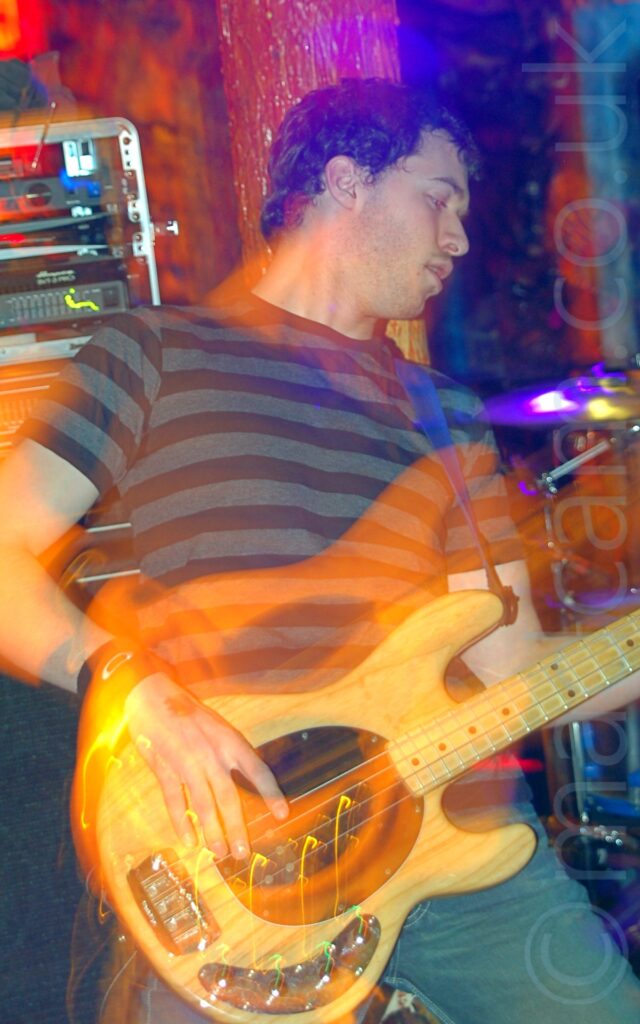 A man with short dark hair, wearing a black and grey striped t-shirt and jeans, playing a varnished wood bass guitar. He is leaning backwards, looking off to his left. A rack of amplifiers in a box is on the left of the background, with an organic-looking orange and yellow thick pole behind his head, and part of a drum kit on the right. A long exposure and his movements have combined with the stage lights to create blooms of orange lights around him and his instrument.
