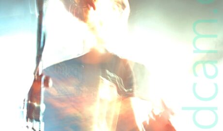 Long exposure shot of a man with short dark hair and wearing a black t-shirt with a brown image filling the chest, and a blue pair of jeans, playing a red electric guitar. The long exposure and the bright stage lights have washed out much of the stage area, while a snap zoom and his movement have left his head dis-jointed from the rest of his body. A microphone on a stand in front of him reflects the stage lights. Black walls and ceiling complete the image.
