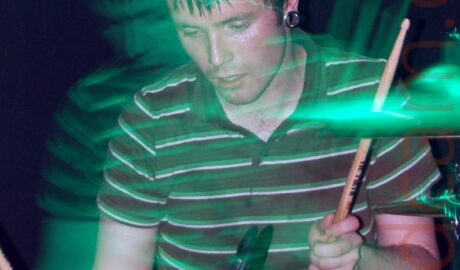A man with short dark hair, metal tunnels in his ear lobes, and wearing a green and white striped polo shirt, sitting behind a red drum kit, with a drum stick held in each hand, held aloft. A flash freezes him in place, but a long exposure and green stage lighting captures his movement (and the movement of the camera) as green secondary images towards the left. A microphone is coming into frame in the top right, and black fabric covers the wall in the background.