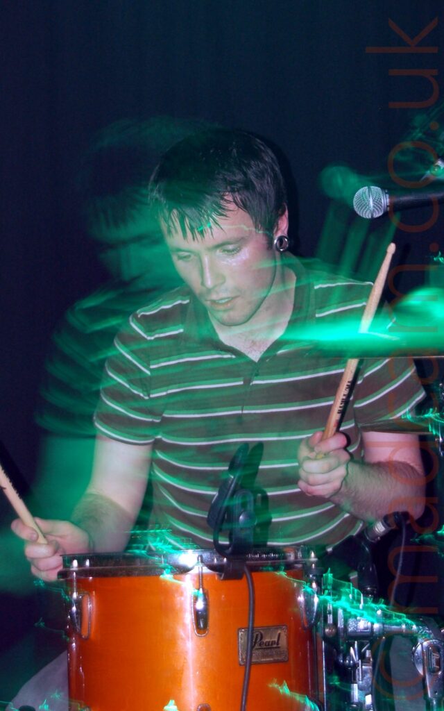 A man with short dark hair, metal tunnels in his ear lobes, and wearing a green and white striped polo shirt, sitting behind a red drum kit, with a drum stick held in each hand, held aloft. A flash freezes him in place, but a long exposure and green stage lighting captures his movement (and the movement of the camera) as green secondary images towards the left. A microphone is coming into frame in the top right, and black fabric covers the wall in the background.