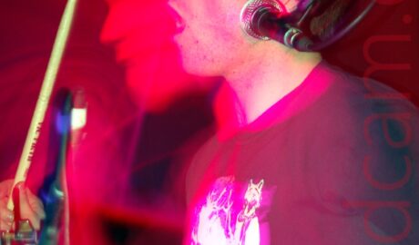 Side view of a man with short dark hair and wearing a black t-shirt with an indistinct white logo. He is sitting behind a drum kit, with a drum stick in each hand, the right stick raised in the air, and is facing to the left of the frame, with his mouth open. There is a microphone hanging down on a stand on the right of the frame, but he isn't currently using it. A flash has frozen him in place, while a long exposure and his movement have created a secondary image of his face shrouded in blooms of red and purple, with red light flooding the black background.