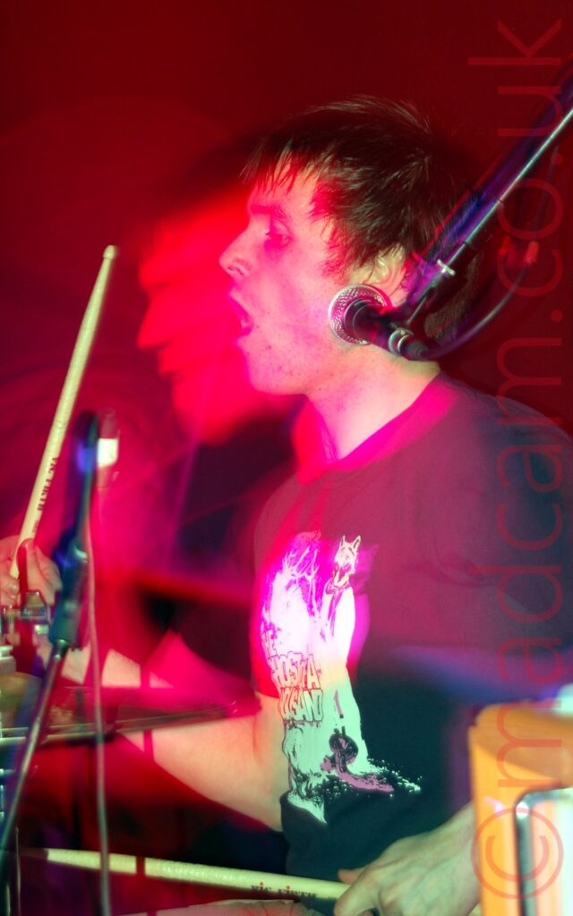 Side view of a man with short dark hair and wearing a black t-shirt with an indistinct white logo. He is sitting behind a drum kit, with a drum stick in each hand, the right stick raised in the air, and is facing to the left of the frame, with his mouth open. There is a microphone hanging down on a stand on the right of the frame, but he isn't currently using it. A flash has frozen him in place, while a long exposure and his movement have created a secondary image of his face shrouded in blooms of red and purple, with red light flooding the black background.