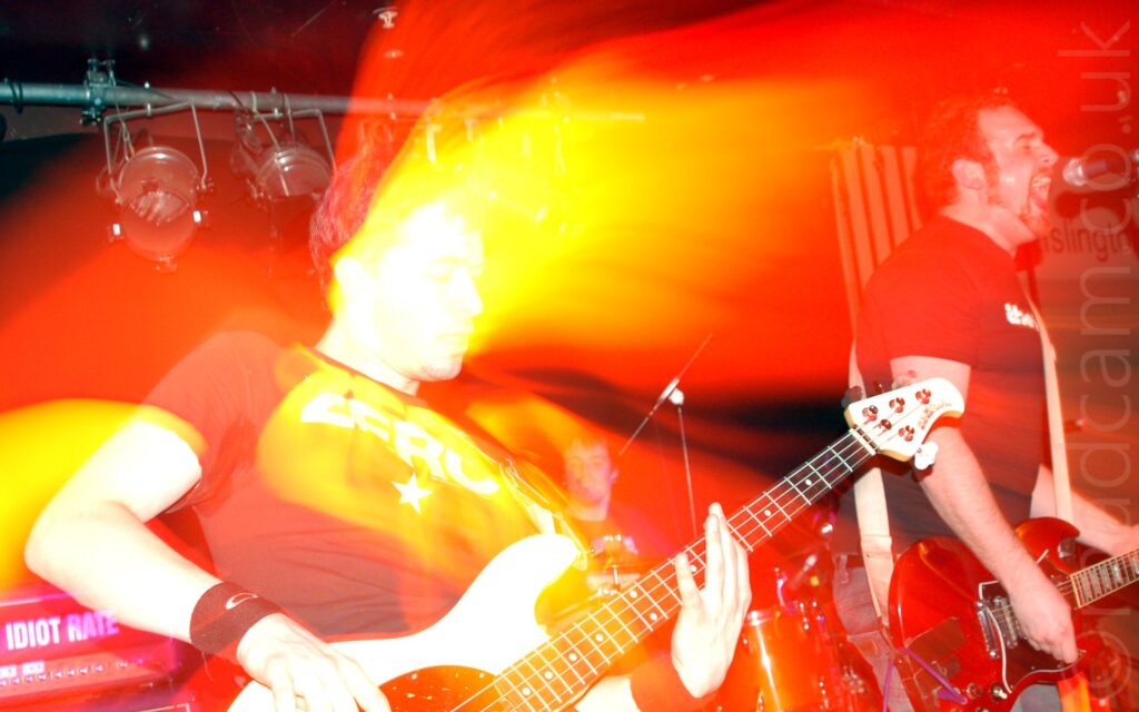 Two men performing on a stage as part of a band. On the left is a man with short dark hair, wearing a black t-shirt, leaning back as he plays a white bass guitar. On the right is a man with short dark hair and a short beard, wearing a black t-shirt, and playing a red electric guitar. A speaker stack with the words "Idiot Rate" on the front is in the bottom left corner, and stage light mounted on a metal pole stretches across the top of the frame. A 3rd member of the band is visible in the background, sitting behind a drum kit. Blooms of red light cover most of the frame, with blooms of white and yellow light surround the bassist, especially his face.