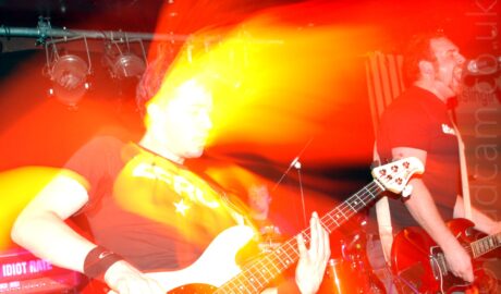 Two men performing on a stage as part of a band. On the left is a man with short dark hair, wearing a black t-shirt, leaning back as he plays a white bass guitar. On the right is a man with short dark hair and a short beard, wearing a black t-shirt, and playing a red electric guitar. A speaker stack with the words "Idiot Rate" on the front is in the bottom left corner, and stage light mounted on a metal pole stretches across the top of the frame. A 3rd member of the band is visible in the background, sitting behind a drum kit. Blooms of red light cover most of the frame, with blooms of white and yellow light surround the bassist, especially his face.