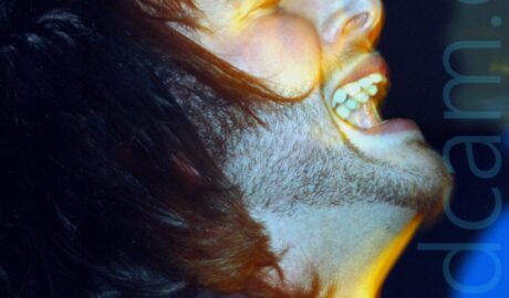 Close-up side view of a man with long dark hair and a short beard, wearing a dark jacket and a scarf knotted around his neck, with his head thrown back and his mouth open, with his eyes closed. Blue and yellow lights bloom across his face and the walls in the background.