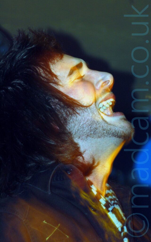 Close-up side view of a man with long dark hair and a short beard, wearing a dark jacket and a scarf knotted around his neck, with his head thrown back and his mouth open, with his eyes closed. Blue and yellow lights bloom across his face and the walls in the background.