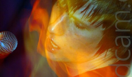 A man with shaggy dark hair, wearing a dark t-shirt, with a light-coloured guitar strap over his left shoulder as he leans backwards towards the camera, with a microphone coming into the frame from the left, beside his face. A long exposure combined with his movement and the stage lights leaves light trails and secondary images streaks of light across the frame.