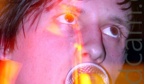 Close-up of the face of a man with short dark hair, his face hidden behind a microphone as he sings, his eyes looking off to the top right of the frame. A long exposure and the stage lights create blooms of yellow and red light, while a snap zoom creates a secondary, almost full length image of him standing behind the mic stand, bathed in yellow light, and holding his guitar in his hands.