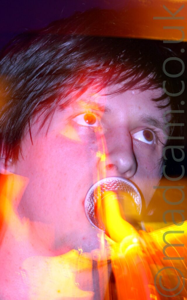 Close-up of the face of a man with short dark hair, his face hidden behind a microphone as he sings, his eyes looking off to the top right of the frame. A long exposure and the stage lights create blooms of yellow and red light, while a snap zoom creates a secondary, almost full length image of him standing behind the mic stand, bathed in yellow light, and holding his guitar in his hands.