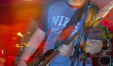 A man with ear-length blond-ish hair, wearing a light blue t-shirt with dark blue sleeves and a large pale blue "Nike" logo emblazoned across the chest, and a blue pair of jeans, playing a black and white bass guitar on a stage as part of a band. There is a microphone on a stand in front of him, but he isn't using it, and in fact it seems to be pointing the wrong way for him. A long exposure and his movement has created blooms or red, orange, and purple lights around him, as various parts of him are illuminated by the stage lights, yet a flash has also frozen him in place. Black walls fill the background, with a yellow light shining brightly on one of the rear walls.
