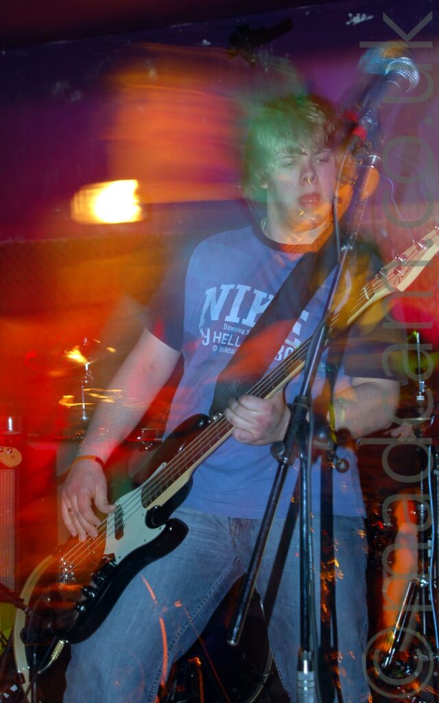 A man with ear-length blond-ish hair, wearing a light blue t-shirt with dark blue sleeves and a large pale blue "Nike" logo emblazoned across the chest, and a blue pair of jeans, playing a black and white bass guitar on a stage as part of a band. There is a microphone on a stand in front of him, but he isn't using it, and in fact it seems to be pointing the wrong way for him. A long exposure and his movement has created blooms or red, orange, and purple lights around him, as various parts of him are illuminated by the stage lights, yet a flash has also frozen him in place. Black walls fill the background, with a yellow light shining brightly on one of the rear walls.