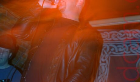 A man with a shaven head and a goatee beard, wearing a black t-shirt and a leather jacket, and blue jeans, singing into a microphone held in his right hand, it's lead held in his left hand. A man wearing a blue t-shirt and a black hat can be seen playing a guitar in the background in the bottom left. A blue drape with grey Celtic knotwork around the edges hangs on the wall in the background on the right. A long exposure and a snap zoom has created a secondary close-up image of the singers head, almost lost in a bloom of red light.