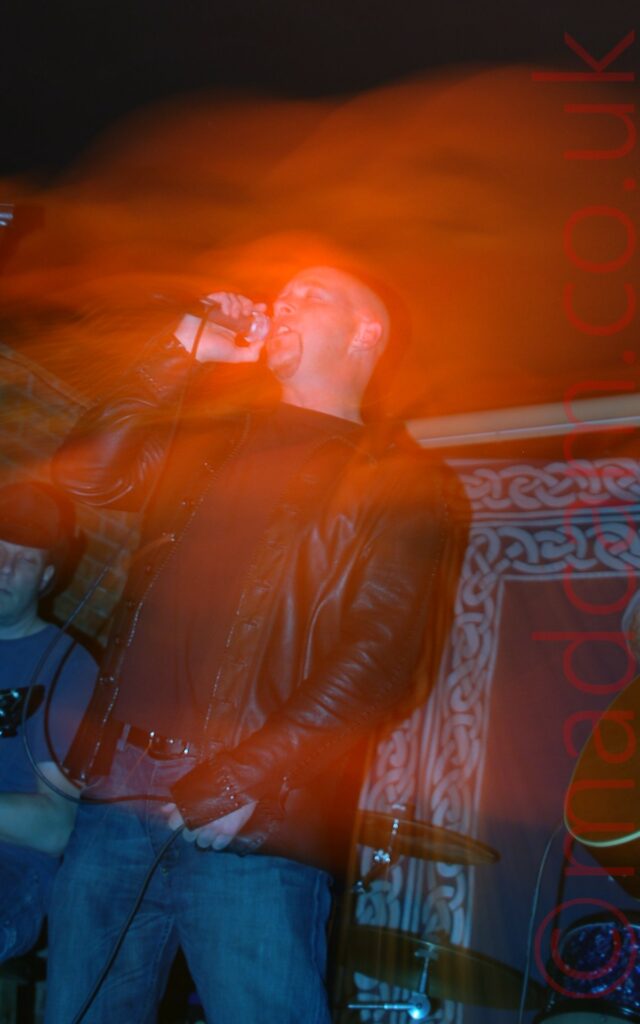 A man with a shaven head and a goatee beard, wearing a black t-shirt and a leather jacket, and blue jeans, singing into a microphone held in his right hand, it's lead held in his left hand. A man wearing a blue t-shirt and a black hat can be seen playing a guitar in the background in the bottom left. A blue drape with grey Celtic knotwork around the edges hangs on the wall in the background on the right. A long exposure and a snap zoom has created a secondary close-up image of the singers head, almost lost in a bloom of red light.