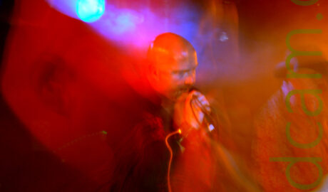 A man with a shaved head, wearing a black shirt with an intricate red floral pattern on the sides, holding a microphone to his mouth with his right hand, with the cord wrapped around his right wrist, leaning over slightly as he sings. A long exposure and a snap zoom has created a secondary close-up image of his head, superimposed on the main shot, with blooms of orange light on the right of the frame, and blue on the bottom left. Black walls and a pair of blue stage lights complete the image.