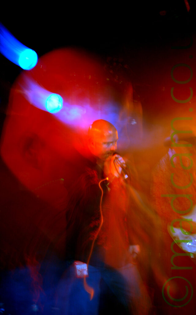 A man with a shaved head, wearing a black shirt with an intricate red floral pattern on the sides, holding a microphone to his mouth with his right hand, with the cord wrapped around his right wrist, leaning over slightly as he sings. A long exposure and a snap zoom has created a secondary close-up image of his head, superimposed on the main shot, with blooms of orange light on the right of the frame, and blue on the bottom left. Black walls and a pair of blue stage lights complete the image.