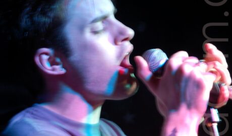 A man with short dark hair singing in to a microphone clasped in both hands, with his eyes shut. He has spiky brown hair, and a tattoo of an black love heart overlaid with a red skull on his right forearm. A long exposure and purple stage lights leaves some artifacts and streaks of light around his hands and face. Black walls fill the background.