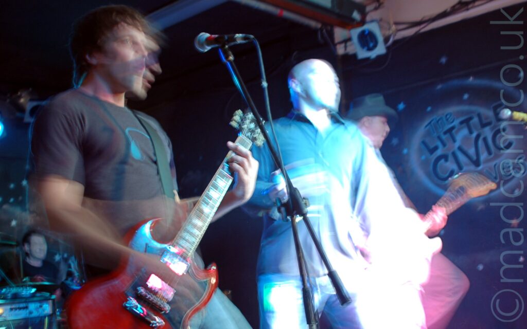 Three men on a stage performing as part of a band. On the left is a man with shaggy dark hair, wearing a grey t-shirt with a blue headphones logo on the chest, and a blue pair of jeans, playing a red electric guitar. In the middle is a man with a shaven head, wearing a dark coloured shirt and blue jeans, holding a microphone in his right hand. On the right is a man wearing a cowboy hat on his head, and playing a an electric guitar, with most of the rest of him hidden behind the middle man. A long exposure and their movements has caught lots of movement, in some cases just blurred parts of the image, in others creating secondary images. In the background, the bands drummer can be seen in the bottom left corner, with black walls filling the rest of the frame.