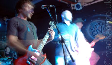 Three men on a stage performing as part of a band. On the left is a man with shaggy dark hair, wearing a grey t-shirt with a blue headphones logo on the chest, and a blue pair of jeans, playing a red electric guitar. In the middle is a man with a shaven head, wearing a dark coloured shirt and blue jeans, holding a microphone in his right hand. On the right is a man wearing a cowboy hat on his head, and playing a an electric guitar, with most of the rest of him hidden behind the middle man. A long exposure and their movements has caught lots of movement, in some cases just blurred parts of the image, in others creating secondary images. In the background, the bands drummer can be seen in the bottom left corner, with black walls filling the rest of the frame.