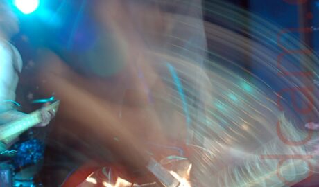Long exposure action shot of a man with shot dark hair, wearing a grey t-shirt and faded blue jeans, facing to the right as he plays a red guitar. Streaks of light and secondary images show his movement. A person playing another guitar can be seen peeking into frame in the background on the left. Blue walls fill the rest of the background, with a single blue stage light shining towards the rear.
