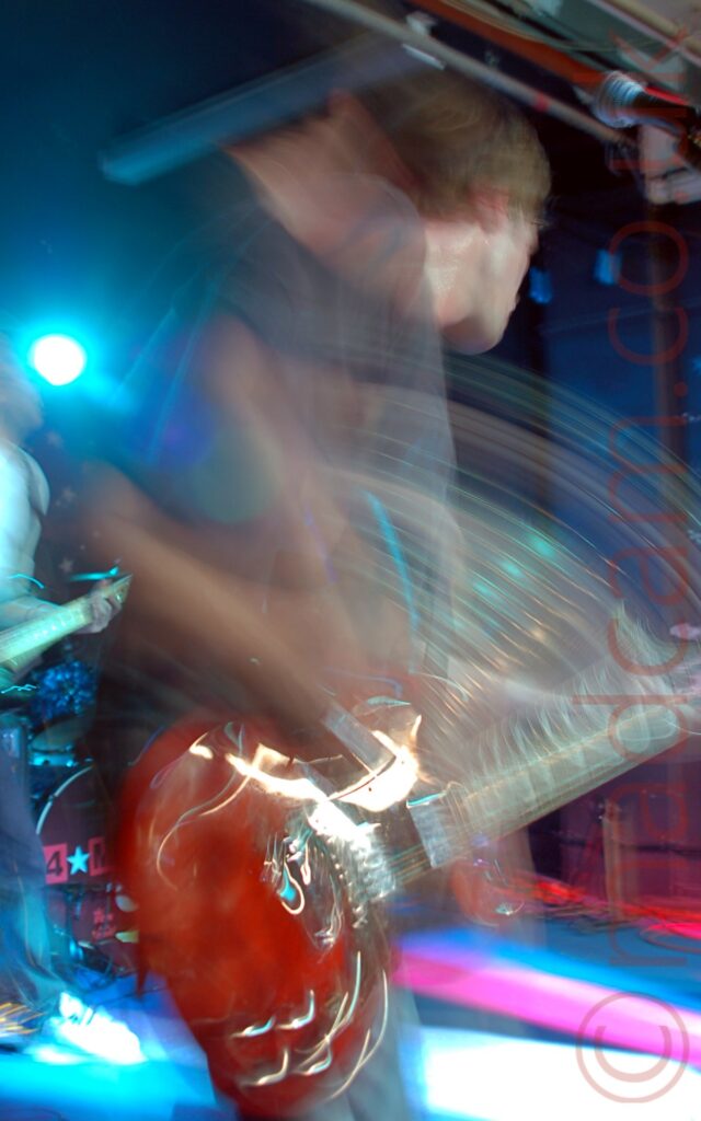 Long exposure action shot of a man with shot dark hair, wearing a grey t-shirt and faded blue jeans, facing to the right as he plays a red guitar. Streaks of light and secondary images show his movement. A person playing another guitar can be seen peeking into frame in the background on the left. Blue walls fill the rest of the background, with a single blue stage light shining towards the rear.