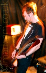 A Man with short dark hair, wearing a black and white t-shirt that resembles a tuxedo with a white shirt and bow tie, standing facing the left of the frame while playing a brown bass guitar. A flash from the camera has frozen him in place, while a long exposure, his movements, and the muted stage lights have left yellow and orange secondary images around him. An amplifier with a weird looking box on top is in the bottom left corner, with textiles draped over the walls in the background.