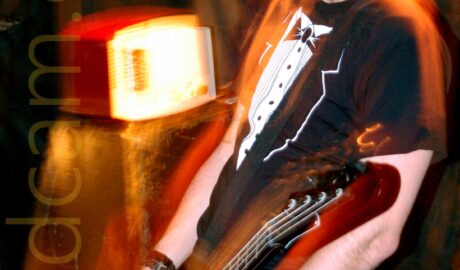 A Man with short dark hair, wearing a black and white t-shirt that resembles a tuxedo with a white shirt and bow tie, standing facing the left of the frame while playing a brown bass guitar. A flash from the camera has frozen him in place, while a long exposure, his movements, and the muted stage lights have left yellow and orange secondary images around him. An amplifier with a weird looking box on top is in the bottom left corner, with textiles draped over the walls in the background.