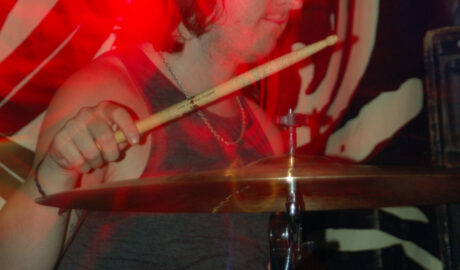 A man with shoulder-length long dark hair, wearing a grey vest top, sitting behind a black and chrome drumkit, with a drumstick in each hand, the right hand raised above a cymbal, the left hand low down over a brown snare drum. A highly decorated black and white drape covers the wall behind him. Red blooms of light seem to congregate on the back of his neck.