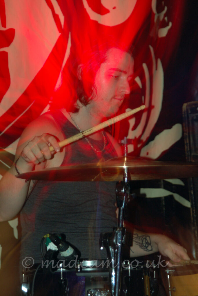 A man with shoulder-length long dark hair, wearing a grey vest top, sitting behind a black and chrome drumkit, with a drumstick in each hand, the right hand raised above a cymbal, the left hand low down over a brown snare drum. A highly decorated black and white drape covers the wall behind him. Red blooms of light seem to congregate on the back of his neck.