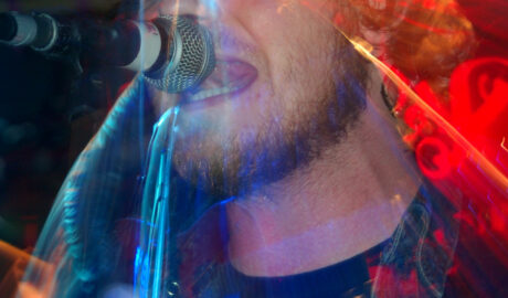 Close-up of the head and shoulders of a man with dark hair and a beard, wearing a black t-shirt with a white, partial text logo on the front, and a blue denim jacket, singing into a microphone on a stand. A long exposure and a snap zoom results in a secondary, smaller image of him, linked to the original with streaks of red, blue, and orange light, showing part of his acoustic guitar, and some of his movements. Black walls fill the background, with blooms of red and orange light on the upper right of the frame.