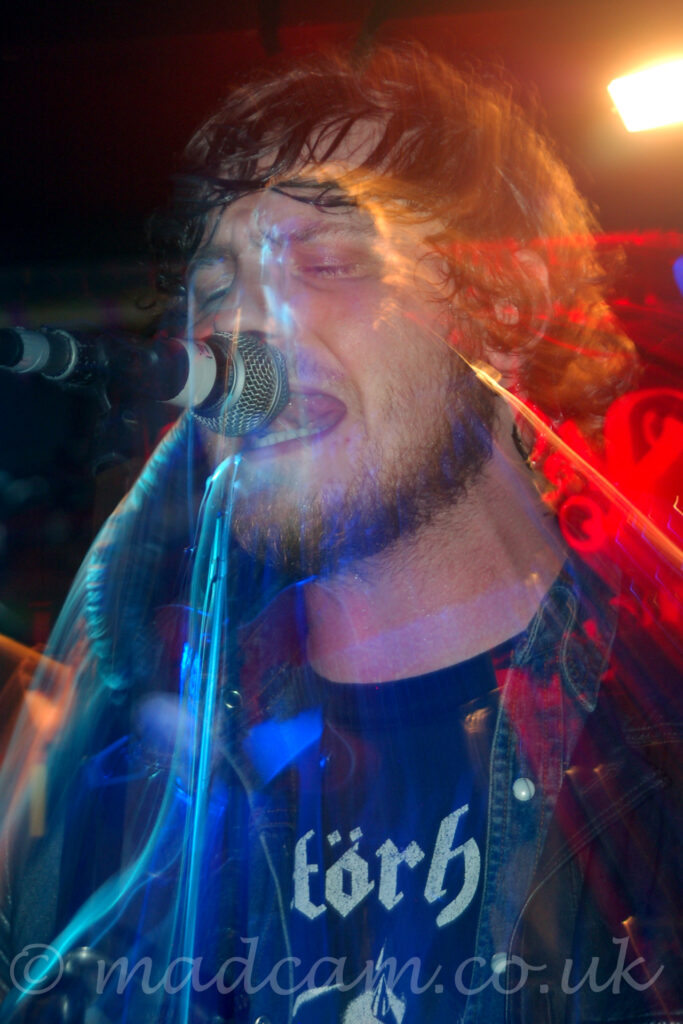 Close-up of the head and shoulders of a man with dark hair and a beard, wearing a black t-shirt with a white, partial text logo on the front, and a blue denim jacket, singing into a microphone on a stand. A long exposure and a snap zoom results in a secondary, smaller image of him, linked to the original with streaks of red, blue, and orange light, showing part of his acoustic guitar, and some of his movements. Black walls fill the background, with blooms of red and orange light on the upper right of the frame.