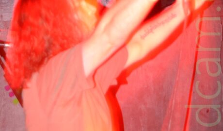 A man with shoulder-length, slightly wavy hair, wearing a grey t-shirt and holding a microphone in his right hand, standing facing to the right, his arms raised in the air in front of him. The stage lights have painted him in various shades of red and orange, while a long exposure and his natural movement have left streaks of light and secondary images all around him, especially around his head and arms. An orange light stares down the lens in the top left corner, while black walls fill the rest of the background.