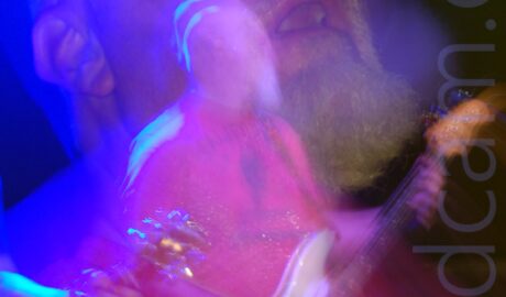 Somewhat blurry shot of a man with a shaven head and white goatee beard, wearing a red t-shirt with dark blue "California" text across the chest, playing a white bass guitar while looking off to his left. The head of an electric guitar being played by someone else is wandering into the bottom left corner of the frame. A long exposure and a snap zoom has created a secondary close-up image of this man's head, tilted slightly back, with his eyes open, a bloom of blue light around his head from the stage lights. A blue and a white light are visible at the top of the frame, with black walls filling the rest.