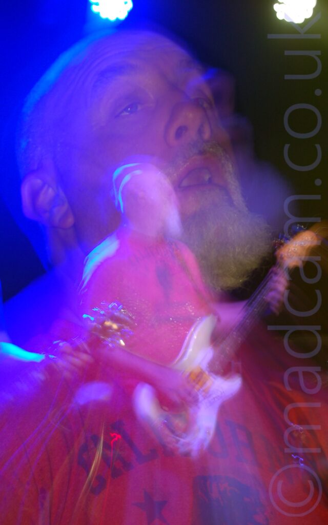 Somewhat blurry shot of a man with a shaven head and white goatee beard, wearing a red t-shirt with dark blue "California" text across the chest, playing a white bass guitar while looking off to his left. The head of an electric guitar being played by someone else is wandering into the bottom left corner of the frame. A long exposure and a snap zoom has created a secondary close-up image of this man's head, tilted slightly back, with his eyes open, a bloom of blue light around his head from the stage lights. A blue and a white light are visible at the top of the frame, with black walls filling the rest.