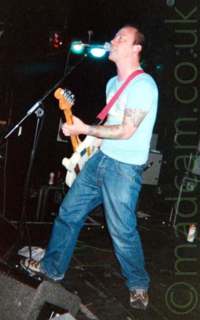 A man with short brown hair, wearing a light blue t-shirt and a partially faded blue pair of jeans, playing a white electric guitar as he sings into a microphone mounted on a stand in front of him. A grey wedge monitor is on the floor in the bottom left corner, with black walls and ceiling filling the background.