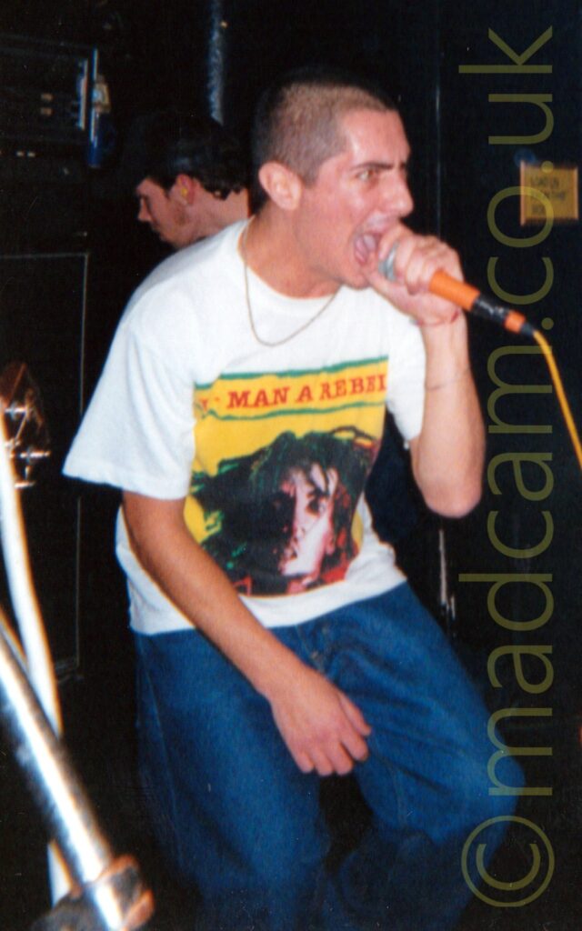 A man with a dark buzz cut hairdo, holding an orange microphone to his mouth with his left hand as he sings. He is wearing a white t-shirt, with a large yellow square filling the chest, with the red text "Every man a rebel" at the top, and the head of a man with long, black dreadlocks at the bottom. He is also wearing a blue pair of jeans. Part of a microphone stand and the head of a guitar is visible on the left of the frame, while a man can be seen leaning in to the side of the stage in the background on the right. Black walls fill the rest of the frame.