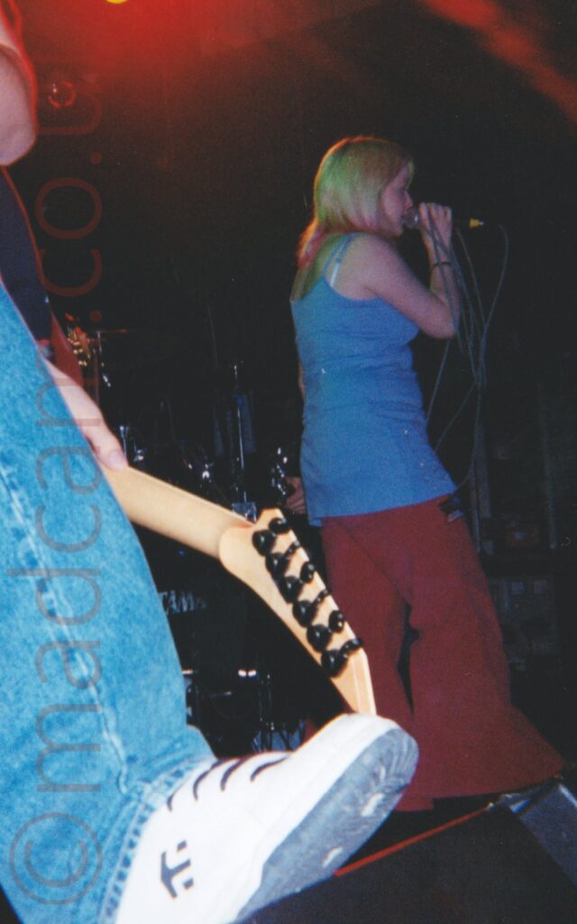 A woman facing to the right, with long blonde hair, wearing a blue strappy top and red baggy trousers, holding a microphone in her right hand to her mouth as she sings. The grey cable on the mic is wrapped loosely wrapped around her hand, most loops extended well past her elbow. In the foreground, the lower left leg and foot of someone with their foot rested on a floor-mounted monitor speaker, clad in a blue pair of jeans and white trainers, with the wooden head of an electric guitar held low down in his left hand. Black walls fill most of the background, with a red stage light illuminating the top of the frame.
