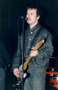 A man with short dark hair, wearing a buttoned-up dark blue jacket and jeans, playing a black and white electric guitar while singing into a microphone on a stand. Another person is almost in the frame on the left, with a red and chrome drumkit in the background on the right. Black walls fill the rest of the frame.