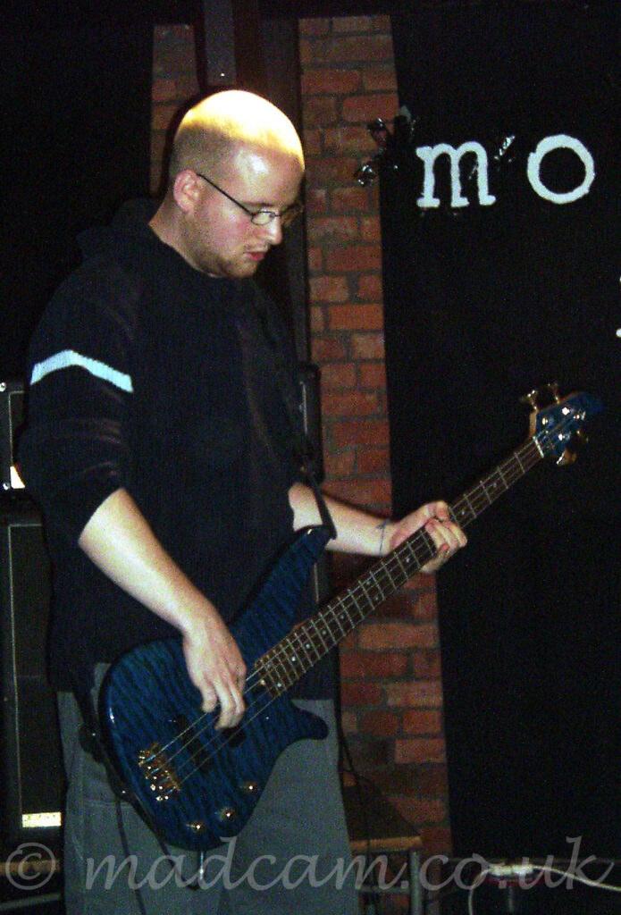 A man with a shaven head, and glasses on his face, wearing a black jumper with the sleeves rolled up, playing a dark blue bass guitar while looking down at the floor. Brown brick walls, partially covered with black drapes, fill the background.