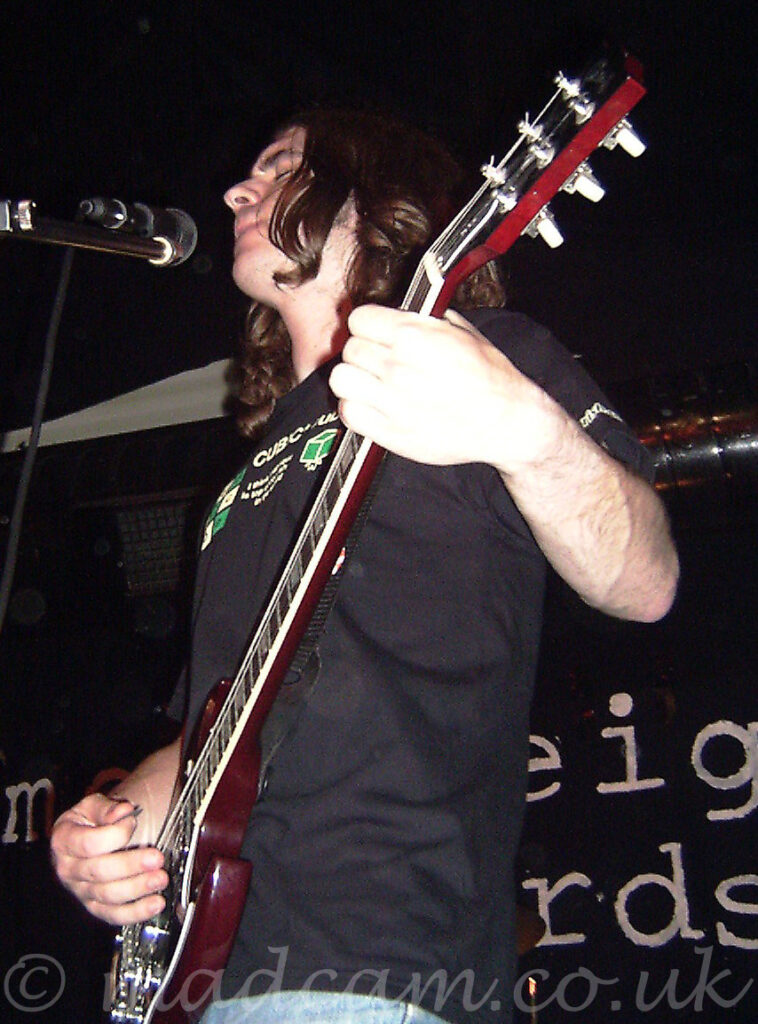 A man with long brown hair hanging to his shoulders, wearing a black t-shirt with an indistinct white and green logo across the chest, and playing a black electric guitar. He is leaning back slightly, lips and eyes closed, seemingly lost in his own world. A microphone on a stand is in front of him, on the left of the frame, but he isn't using it in any way. Black walls and ceiling fill the background.