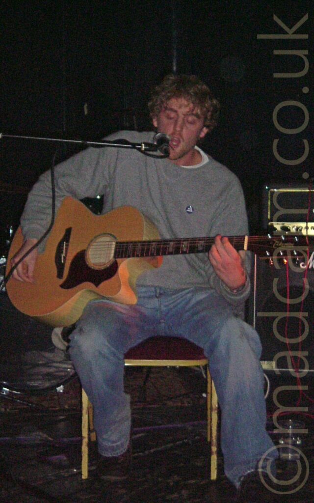 A man with fairly short, slightly curly , light coloured hair wearing a grey jumper over a white t-shirt and a blue pair of jeans, sitting on a chair and playing a brown acoustic guitar while singing into a microphone on a stand in front of him. A small stack of amplifiers is sitting on the floor behind him and to his left, with black walls filling the rest of the frame.
