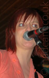 A woman with chin length red hair, wearing a peach-coloured top, singing into a microphone on a stand that covers her mouth, her eyes tilted slightly to look off to her left. Black and brown ceiling and walls fill the rest of the frame.