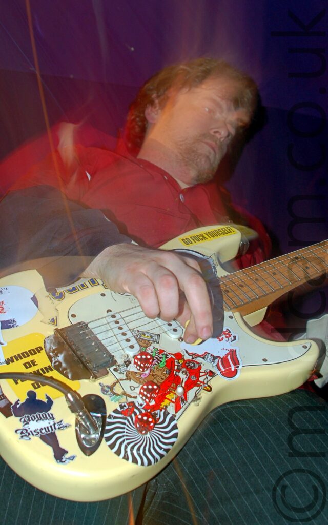 A low-down, long-exposure shot of a man with brown hair, wearing a red shirt over a black jumper, and black trousers with grey pinstripes, and playing a yellow electric guitar, covered in many multi-coloured stickers. A single trail of red light runs from the top left corner to the bottom middle of the frame, with more streaks around his head. Purple walls and ceiling fill the background.