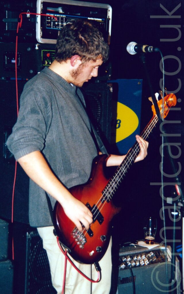 A man with short dark brown hair and sideburns, wearing a grey jumper and grey trousers, playing a black and brown bass guitar, staring down at his left hand, as he faces right. A microphone on a stand is in front of him, on the right of the frame. A stack of amplifiers on top of a speaker cabinet is behind him, as well as a bass amplifier on the floor in the bottom right corner, with a partially drunk pint on top. Black walls and ceiling fill the rest of the frame.