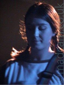 A woman with long dark hair, wearing a white long-sleeved t-shirt with blue piping around the neck and dark blue sleeves, and a rainbow-coloured guitar strap over her left shoulder. She is looking down pensively. The whole frame has a slight blue cast, apart from the back of her head, which is illuminated by a bright white light.