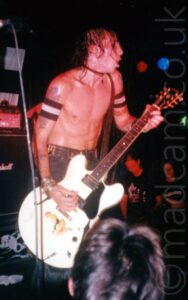 A man with his shirt off, with lank dark hair and wearing a studded belt holding his blue jeans up and playing a large, white, electric guitar, facing to the right of the frame. He is absolutely drenched with sweat. A pair of arm bands are round his biceps, white in the middle, with black stripes at top and bottom. The head of someone in the crowd can be seen right at the bottom of the image. A stack of speakers can be seen in the background on the left of the frame, while two people watch from the side of the stage on the right. Black walls and ceiling fill the rest of the image.