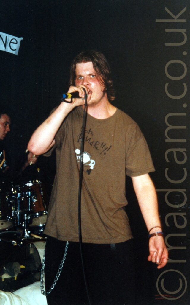 A man with chin-length, rather floppy dark hair, wearing a brown t-shirt with a black text logo and white and grey cartoon style images on the chest, performing on a stage as part of a band, holding a microphone to his mouth as he sings. Another man can be seen sitting behind a black drumkit on the left of the frame, with black walls and ceiling filling the rest of the background.