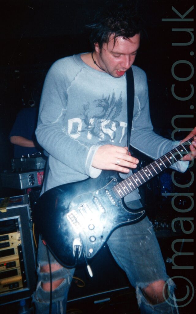 A man playing a guitar on a stage, performing as part of a band. He has slightly spiky black hair, and is wearing a pale blue long-sleeved t-shirt, with the dark blue silhouette of a rearing dragon overlaid with the white word "DIRT" on the chest, and a ripped, faded blue pair of jeans, and playing a black electric guitar attached to a black strap over his left shoulder. A bright light is shining directly on the middle of the guitar. A stack of gold-coloured amplifiers in a black travel case is in the background on the left of the frame, with a man wearing a dark blue t-shirt behind a set of turntables behind that. Black walls and ceiling fill the rest of the frame.