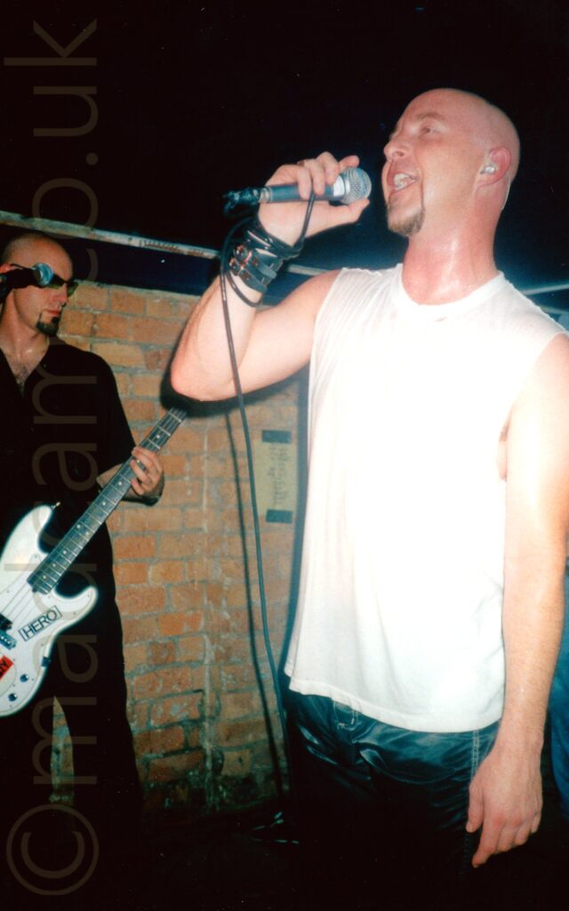 Two men performing on stage as part of a rock band. On the left, almost sneaking out of frame, is a with a shaven head and a black goatee beard, wearing a pair of sunglasses, a black shirt, and black trousers, and playing a grey bass guitar. On the right and nearer to the camera is a shaven-headed man with a brown goatee, wearing a sleeveless white t-shirt and blue jeans, singing in to a microphone held in his right hand, it's lead wrapped around his right wrist. A brown brick wall and black ceiling fills the background.